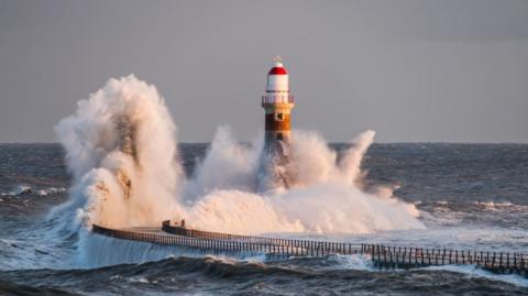 Roker Pier. Large waves are crashing into the pier and it's lighthouse. The pier curves to the right and has railings along each side. The lighthouse has a white and red dome.