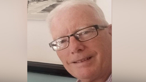 Andrew Avery smiling for the camera. He is wearing glasses. There is a picture on the wall behind him.