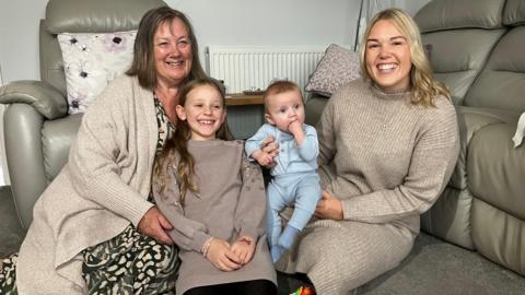 Two grown women in light brown cardigans with one little girl in a brown dress and a baby boy in a blue babygrow are sat on the two women's lap