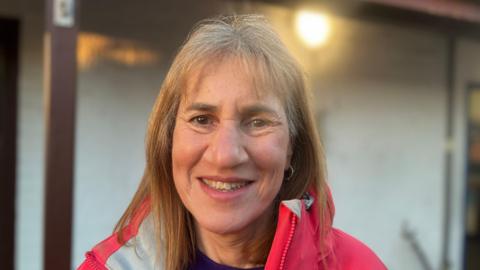 Elaine has long brown hair and is wearing a pink and white ski jacket