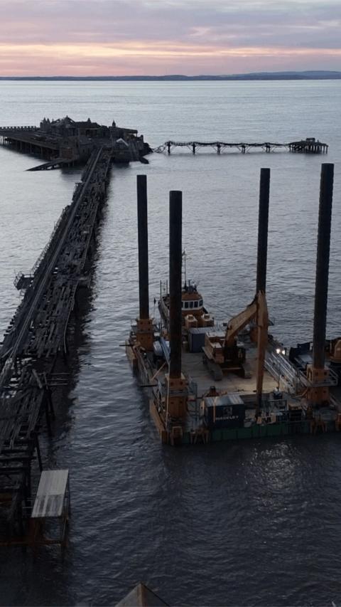 A large specialist barge with four support poles in each corner sits in the water next to Birnbeck Pier at sunset