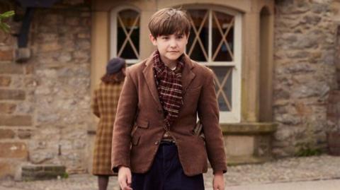 A photograph of Owen on set, wearing a brown jacket and scarf, walking across a square.