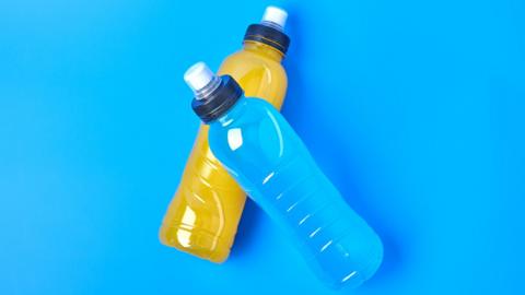 A drinks bottle full of blue liquid, on top of a bottle of orange liquid, on a blue background
