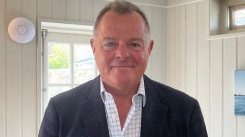 Michael Barnes has short dark hair and is wearing a dark jacket and white checked shirt. He is standing indoors and is looking straight at the camera, smiling.