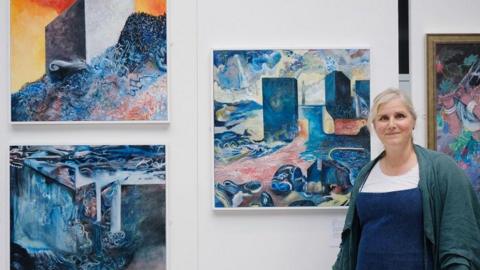 Artist Tara Harris stands next to paintings displayed on a gallery wall.