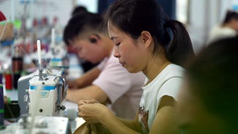 A woman sits at a sewing machine in a factory