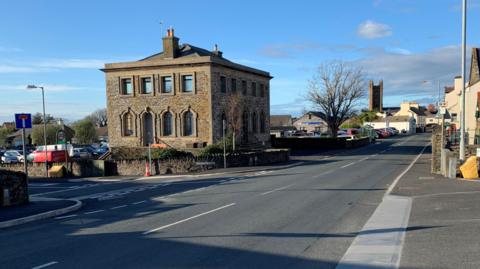 The old bank building on the corner of Station Road in Kirk Michael. It is a two-storey brick building with a wall around it.