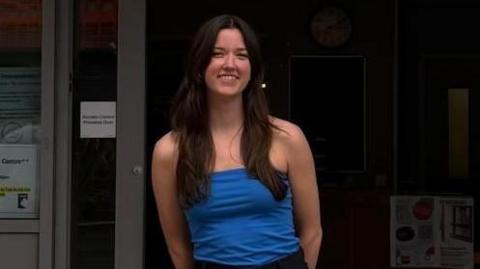 Charlotte Buttercase, who has long brown hair, is smiling at the camera in front of a building with the sign reading: Undergraduate Centre. 