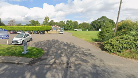 Cars are parked on a parking area with hedges, a pavilion and playing fields in the background.