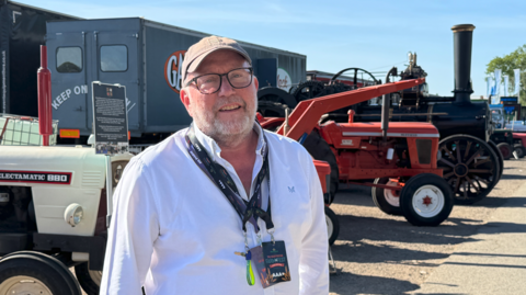 A man wearing a white shirt, a cap, glasses and two lanyards is looking at the camera. Part of some agricultural vehicles are behind him on a sunny day.