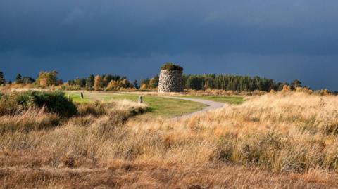 A very dark grey sky above Culloden battlefield. There is rough scrub in the foreground, beyong which is a path and neatly cut grass. A short circular stone tower is in the middle of the picture. 