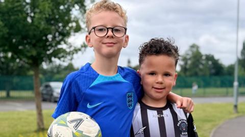 Two children wearing football jerseys pose proudly for the camera. The child on the left holds a football.