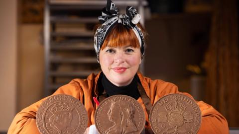 Chocolatier Jen Lindsey-Clark unveils a collection of historical milk chocolate coins which were created to celebrate the opening of Cash Access UK's 200th banking hub.  There are three coins, roughly the size of small dinner plates placed in front of her. She is sitting behind the coins smiling, in an orange coat or jacket.