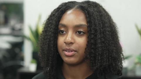 A close-up of Jayzik sitting in front of some house plants wearing a black top. She has a small nose piercing and shoulder-length hair. 