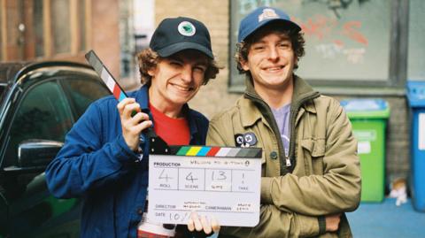 Twin brothers wearing caps smile as they pose with a movie clapperboard