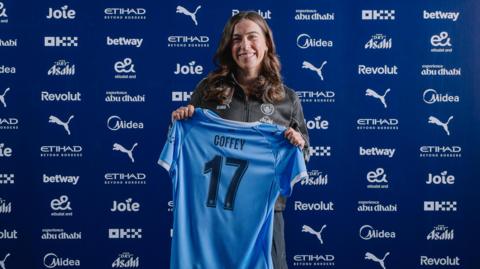 A smiling Sam Coffey holds a blue Manchester City shirt with Coffey 17 on its back, as she stands in front of a wall showing sponsor logos