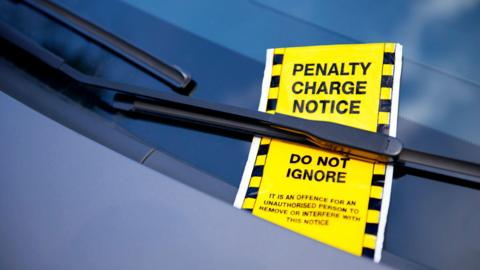 A bright yellow ticket under a car windscreen wiper reads "penalty charge notice do not ignore: it is an offence for an unauthorised person to remove or interfere with this notice".