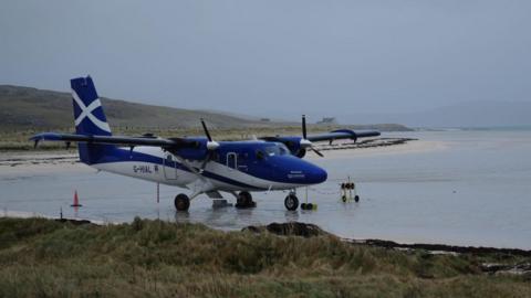 The aircraft is blue and white in colour. It has two propeller engines and is stationary on the sand of Barra's beach airport. It is a grey and chilly day.