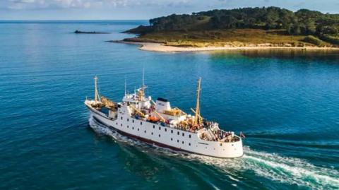 A white ferry crowded with passengers on its decks leaves a white wake as it crosses the blue sea. There is a mass of land in the background with trees, grass and sand on it.