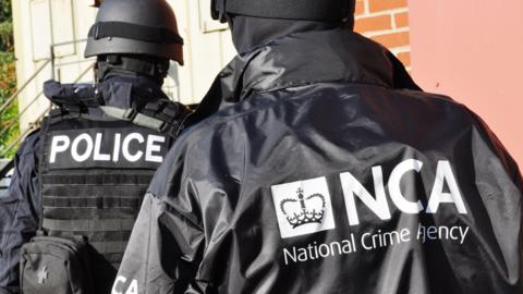 The back of two officers - one wearing a dark police vest and another with a National Crime Agency coat on. They are both wearing helmets. 