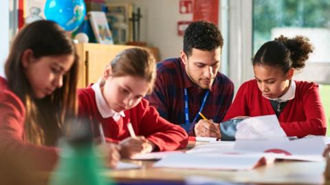 Male teacher sitting at a desk in a classroom with three female pupils who are all working. The teacher is wearing a red and black checked shirt and has a lanyard round his neck. The pupils are wearing red jumpers or cardigans. There are books on the desk and books and a globe behind them on a cabinet