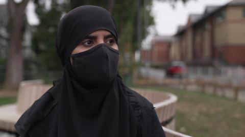 A woman wearing a black hijab and black face mask stands in front of a grassy area of a residential street.