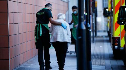 An elderly women being led passed an ambulance by a paramedic wearing a green uniform and an apron. He has his arm around the lady's back. 