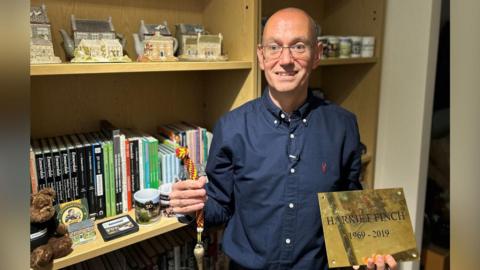 A man holding a yellow and red rope in his right hand with a bell attached to the bottom and a gold plaque in his left with the words HARRIET FINCH 1969-2019 written on it. Behind him is a book shelf with books and mugs on. He is wearing a blue long-sleeve shirt and has a bald head and is also wearing glasses.