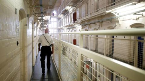 A prison officer - with a white shirt, black trousers and bald head - walks along a row of cells at Barlinnie prison. 