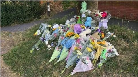 A large pile of bunches of flowers and soft toys left at the base of a tree