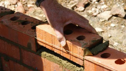 Close up picture of a hand placing a red brick on top of some mortar as a small wall is being built in the sunshine.