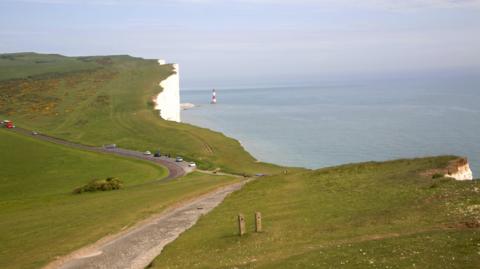 A coastal view of Beachy Head featuring white cliffs, green fields and a striped lighthouse by the sea.