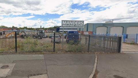 A pavement is in the foreground, with the words Manders industrial estate on a sign behind a fence. Vehicles and a building are behind the sign.