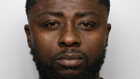 A mugshot of Boachie, photographed from the front against a plain, light‑coloured background. He has short, closely trimmed hair and a beard. He is wearing a dark T‑shirt.
