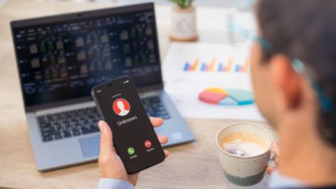 Businessman holding a smartphone with displaying an incoming suspected call from unknown caller on his smartphone, contemplating whether to answer a potential scam call while working at a desk with trading board on a computer screen.