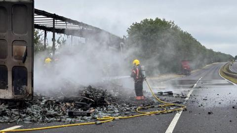 A firefighter wearing a blue and yellow fire suit, holding a yellow hosepipe, putting out a fire on the road. Debris and smoke can be seen on the road.