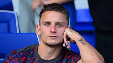 Luke Woolfenden stares ahead with his chin resting on his hand. he has a tattoo along his forearm saying 'maia' and wears a red and blue patterned training t shirt