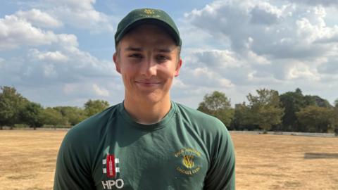 Young man with a green cap on and a green long sleeve top. He is smiling. He is stood on the edge of a cricket field and the grass is parched and sandy coloured. 