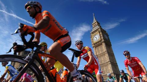 Cyclists in lycra and helmets travel past Big Ben, with blue skies in the background. 