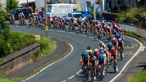 A peloton of cyclists cycling round a corner on a Welsh road over a small stone bridge