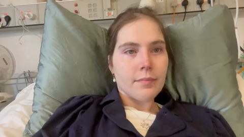 Lily McGarry lying in a hospital bed. They are wearing a dark garment and resting on two pillows. The background includes typical hospital equipment and electrical outlets.

