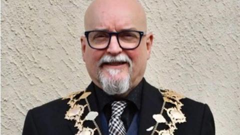 An elderly bald man with black-framed glasses and a white goatee beard smiles at the camera. He is wearing a dark suit, a black shirt and a checkered tie, as well as a chain of office.