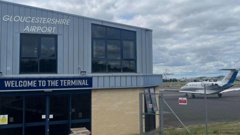 A shot of the Gloucestershire Airport terminal building with a small private jet plane to its right. The building is a small two storey building with the airports name written above and a sign saying Welcome to the Terminal written above the door