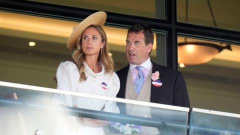 Harriet Sperling and Peter Phillips during day one of Royal Ascot at Ascot Racecourse, Berkshire. She is wearing a white jacket with puffed sleeves and a straw hat which sits at an angle on her long, blonde hair. He is wearing a dark-coloured jacket, a light grey waistcoat, a pink shirt with a white collar and a light blue tie. 