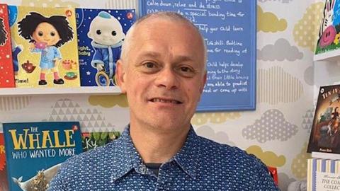 Duncan Cooper has short grey hair. He is wearing a blue and white patterned shirt with a black T-shirt underneath. He is stood in his bookshop with children's books on white shelves behind him.