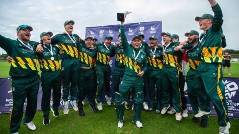 The Guernsey team, dressed in green tracksuits with orange and yellow patterns across the midriff, celebrate reaching the final stage of T20 World Cup qualifying for the first time 
