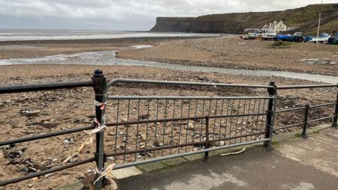 A temporary fence has been attached to the Victorian railing and there are straggly pieces of red and white tape dangling off it. Beyond it is a stony and sandy beach with a channel of shallow water snaking towards the sea. Bots, buildings and cliffs can be seen in the distance.