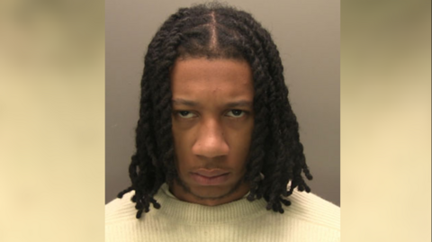 A police custody image of UK rapper Digga D whose name is Rhys Herbert. He has long, braided black hair and dark eyes. He is wearing a cream coloured jumper.