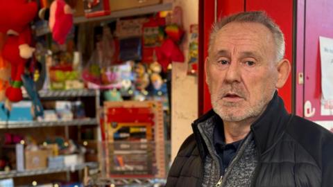 David Taylor with short grey hair and beard wearing a grey zip up jumper and black jacket. He stands in front of a toy stall in a market.