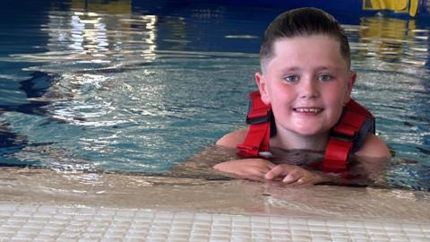 Rory with brown wet hair wearing a red life-jacket is in a swimming pool with his hands resting on the edge. Rory is smiling.